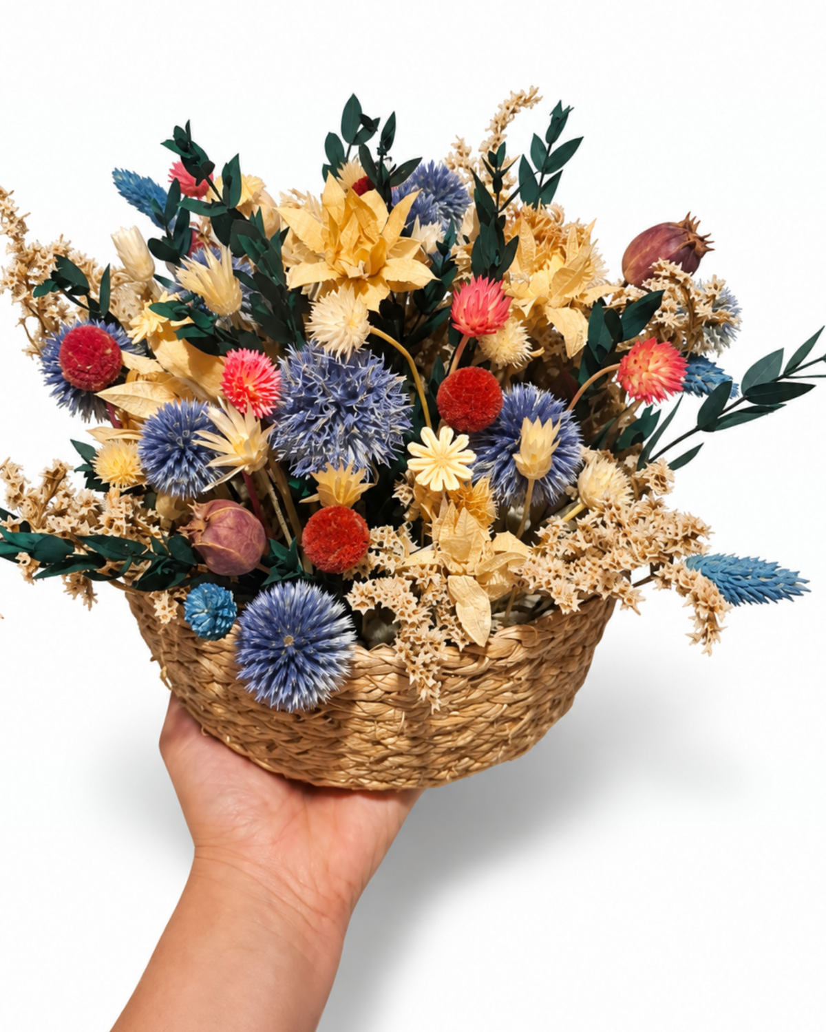 Hand holding a medium woven basket filled with vibrant dried echinops, red craspedia, and safflower blooms.
